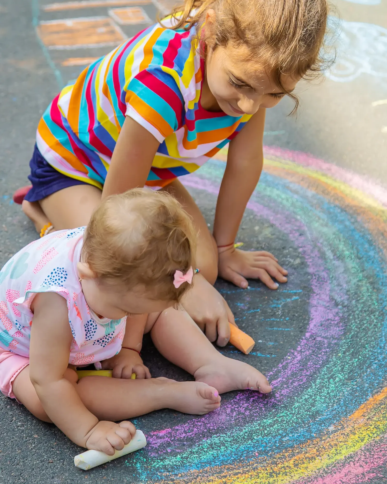 Two young girls kneeling on a sunny driveway, drawing a big rainbow in coloured chalk