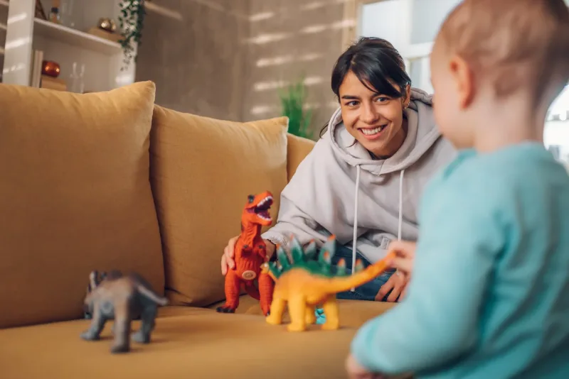 A smiling nanny kneeling at a couch playing toy dinosaurs with a toddler