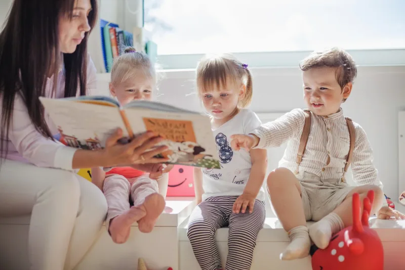 A caregiver reading a picture book to three young children sitting by a sunlit window